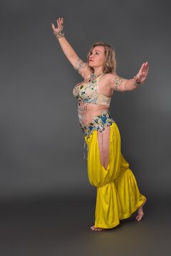 Mid aged adult woman belly dancer with raised and outstretched arms in dancing costume - bra top and yellow harem trousers and she is not wearing any footwear, performing full length, gray background