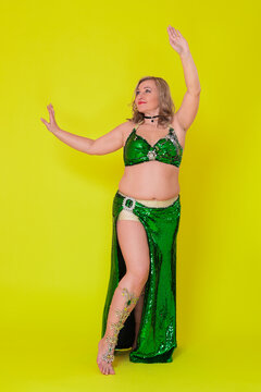 Curvy woman dancer with raised arms dressing in dancing stage costume - green sequined bra top and long skirt with slit and she is not wearing any footwear, performing full length yellow background