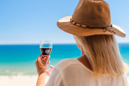 Woman in straw hat holding glass of red wine while enjoying tropical beach and turquoise sea view during luxury summer vacation