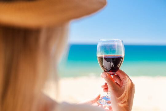 Woman holding glass of red wine with turquoise sea and sandy beach in background during relaxing luxury summer getaway