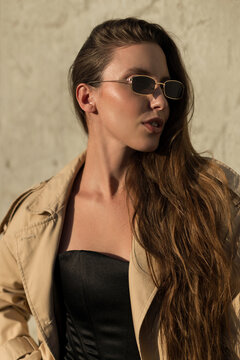 Stylish young woman with long wavy hair, small rectangular sunglasses, and a beige trench coat turned sideways in warm golden sunlight against a concrete wall background. Urban fashion portrait.