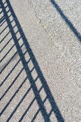 Fototapeta premium A high angle shot of a geometric shadow cast by a metal railing fence on a rough asphalt surface during a sunny day.