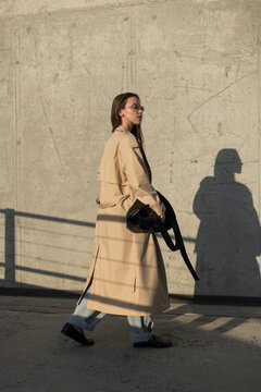 Side view of a stylish young woman walking in a classic beige trench coat and jeans past a concrete wall with strong sunlight and sharp shadows. Modern street fashion concept.