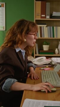 Vertical video of young adult Caucasian woman typing on retro computer keyboard and using mouse at office desk. Clip is ideal for vintage technology or 90s workplace themed marketing campaigns