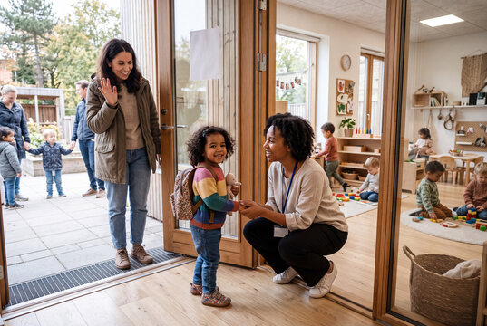 A daycare teacher watches with a smile as a child stands at the door. The parent waves goodbye. Other children can be seen playing inside the classroom