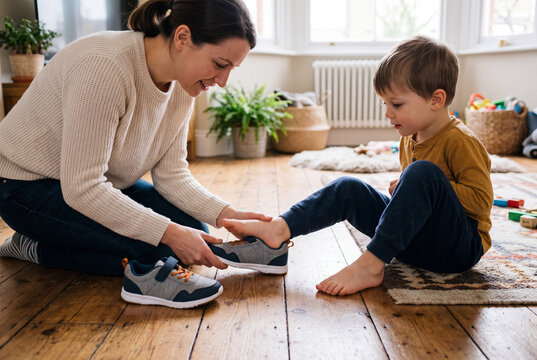Parent sits on the floor with child. They check the fit of new sneakers against the child's foot. The room has natural light and wood floors. Toys are visible in the background