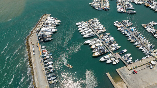 Aerial top view of a crowded marina with rows of docked yachts and boats, where a small boat enters leaving a gentle wake on the clear turquoise sea water.
