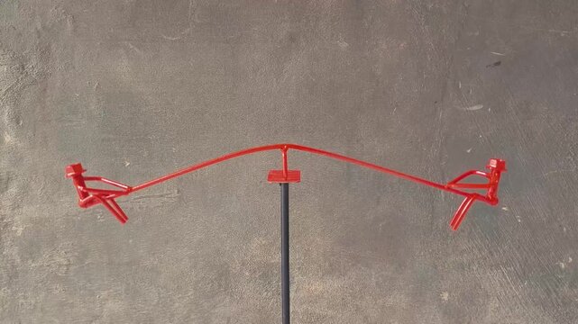 An artistic miniature seesaw, meticulously crafted from bent metal wire and bolts, and painted a vibrant red. The two seats are ingenious representations of small human figures, complete with bolt hea