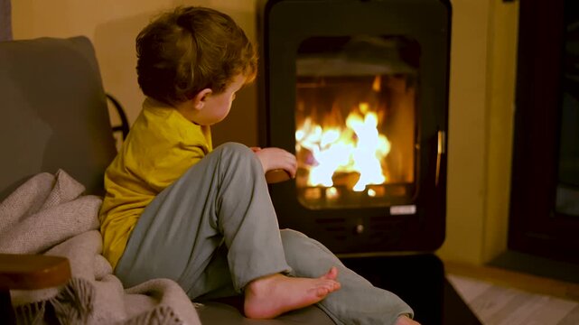 A young child sits mesmerized by dancing flames in a cozy home. The little boy watches the warm light flicker, feeling peaceful and calm in the evening atmosphere.