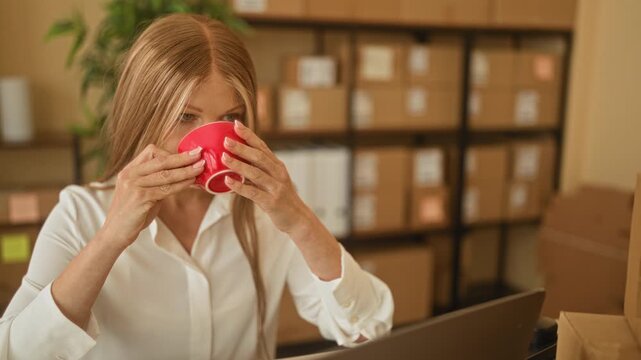 Young woman holding red mug at laptop amid stacked shipping boxes and parcel shelves in a packing building; calm concentration.