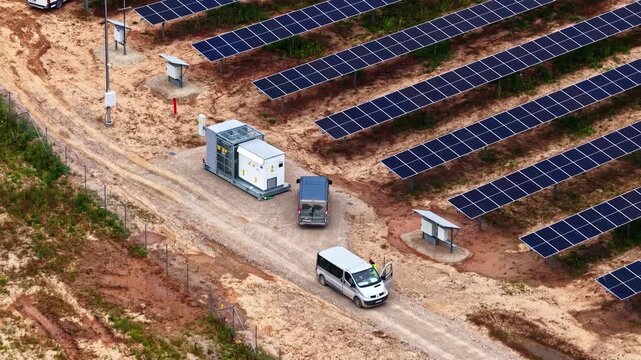 Aerial view of a solar farm with rows of photovoltaic panels, a power substation, and service vehicles parked on a dirt road in Tume, Tukums Municipality, Latvia.