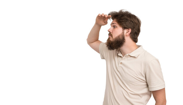 Bearded young man shading his eyes with hand while looking into the distance isolated on white background, searching, vision and future concept.