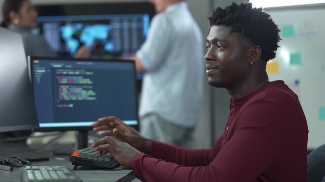 Young African male software engineer writing program code on multiple desktop screens while his colleagues working in the background in a modern office.