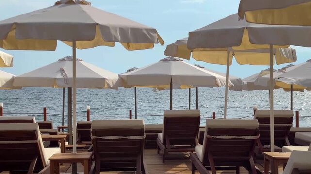 A soothing seaside view from the wooden deck of a luxury resort or beach club. Rows of neat wooden sunbeds are visible beneath cream-colored beach umbrellas swaying gently in the breeze.