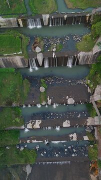Aerial view of Grojogan Watu Purbo waterfalls with cascading water over stone dams and lush riverbanks in Kecamatan Tempel, Daerah Istimewa Yogya, Indonesia.
