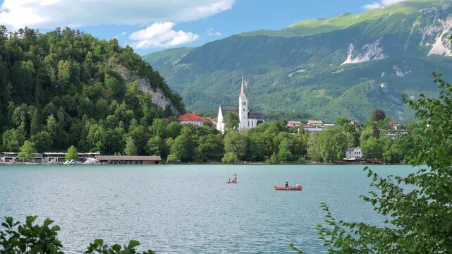 View of Lake Bled in Slovenia with unrecognizable people rowing a boat, St. Martin's Parish Church and the majestic Julian Alps creating a stunning alpine backdrop.