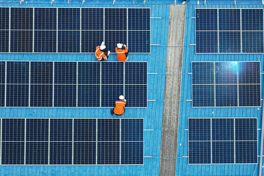 Aerial view of technicians installing solar panels on blue roof. Engineers maintaining solar energy system on a building rooftop. Top down view of workers in orange uniform fixing solar panels. 