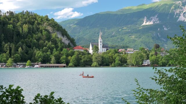 View of Lake Bled in Slovenia with unrecognizable people rowing a boat, St. Martin's Parish Church and the majestic Julian Alps creating a stunning alpine backdrop.