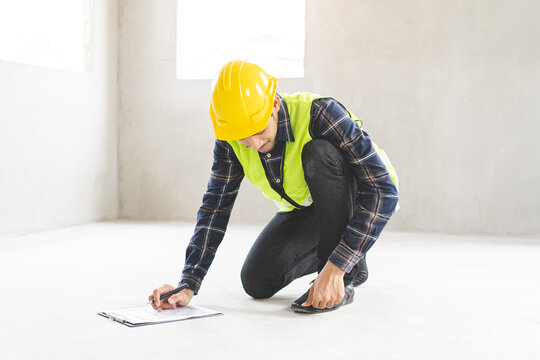 Asian male construction engineer or inspector in safety uniform checking concrete floor quality and writing on clipboard checklist at home building renovation site project.
