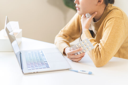 Sick asian young woman holding her sore throat and a blister pack of pills while consulting a doctor online via laptop at home, telemedicine and remote healthcare technology concept.