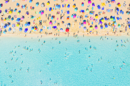 Aerial drone view of people relaxing on a white sand beach under colorful umbrellas. Bright sunny summer vacation scene by turquoise crystal clear sea water.
