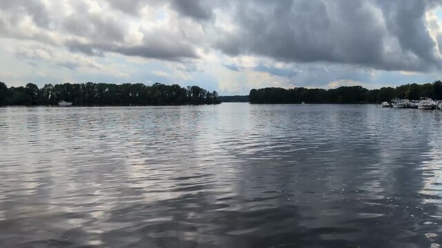 wide view of a calm lake under a dramatic, cloudy sky with gentle water ripples and treelines in the background.