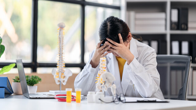 Asian Adult Doctor Feeling Stressed at Desk with Spinal Model Medical Bottles Healthcare Challenges Physician Burnout Concept