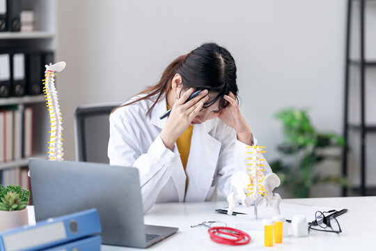 Stressed Asian Woman Doctor with Spinal Model at Desk Feeling Overwhelmed Burnout Exhausted in Clinic Office Spine Pain Healthcare Medical Professional
