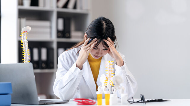 Stressed Asian Female Doctor with Headache at Desk Overwhelmed with Work Discomfort Healthcare Professional Burnout Mental Health Spine Model