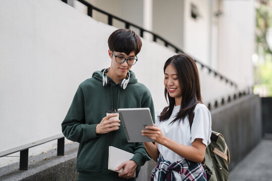 Two young people are standing on a sidewalk, one of them holding a tablet