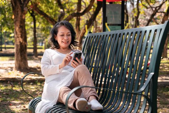 Older Asian Woman Relaxing on Park Bench Using Smartphone for Communication and Connection in Green Scenery Promoting a Peaceful Retirement and Leisure
