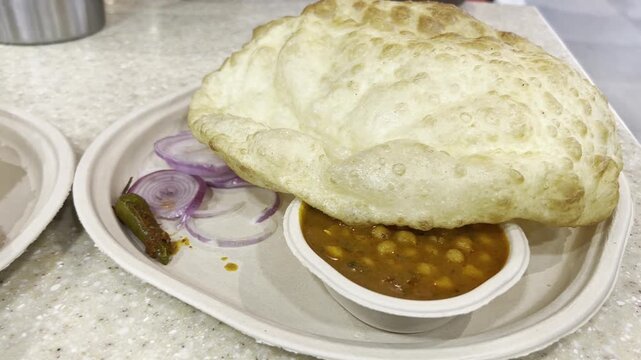 Close-up Full HD footage of fluffy fried bhatura served with spicy chickpea curry, onion slices and green chili pickle on disposable plate, showcasing traditional Indian street food restaurant meal.
