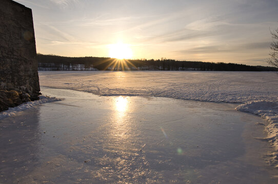 Winter sunset over frozen Lake arr in Dalsland, Sweden; golden sun rays shining behind an old stone ruin on a snowy shore with a peaceful, scenic wilderness atmosphere