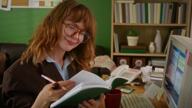 Young adult Caucasian woman with glasses flipping through notebook and working at cluttered retro desk with CRT monitor, tilt up. Perfect for nostalgic corporate training or vintage marketing