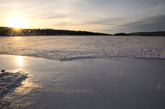 Winter sunset over frozen Lake arr in Dalsland, Sweden; golden sun rays shining behind an old stone ruin on a snowy shore with a peaceful, scenic wilderness atmosphere