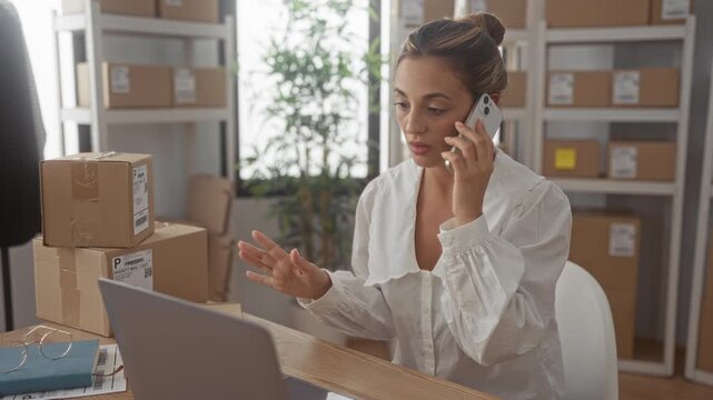 Woman talking on phone while typing at laptop with stacked parcels on desk in building; small business productivity.