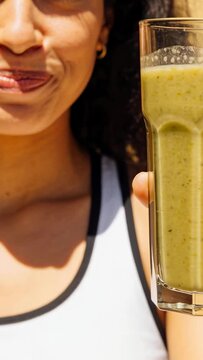 Vertical video: Smiling woman in white top sipping green smoothie to taste on patio as camera moves