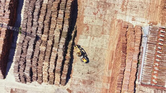 A large facility shows stacks of logs arranged in rows on a dirt surface. A machine operates in the center of the area, moving logs around. The scene is busy with wood processing activity.