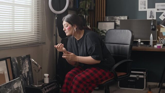 Young adult woman applying hand cream and touching face while looking in mirror at home. Perfect for skincare product advertisements or self-care vlogging content