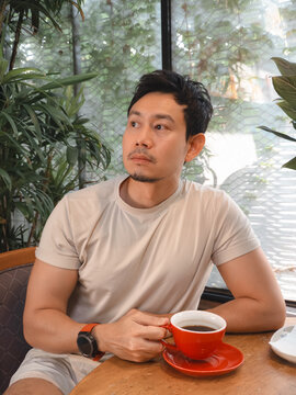 Pensive Asian man sips black coffee at a cozy cafe table by a window.