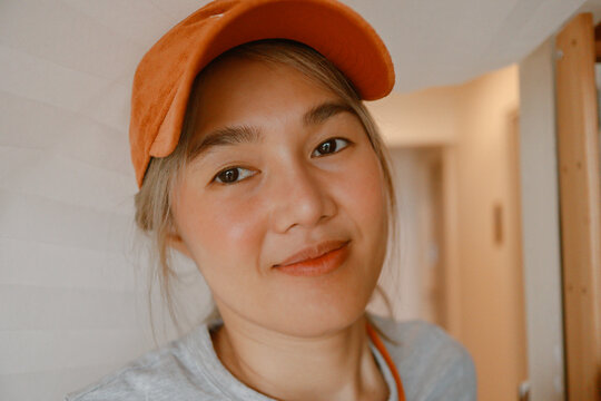 Close-up portrait of a smiling young Asian woman wearing an orange baseball cap indoors.