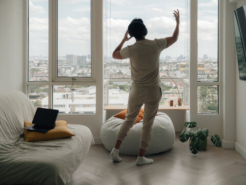 Relaxed man in headphones dancing by a window, enjoying music and city views at home.