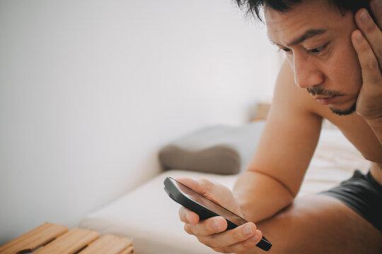 Serious Asian Man Checking His Mobile Phone in Bed, Looking at Screen with Concern