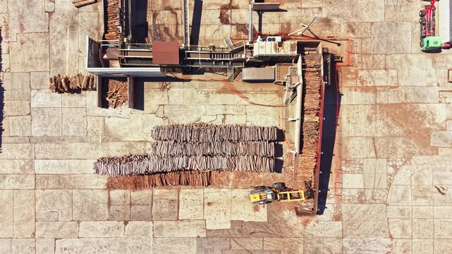 A facility shows different stages of wood processing. Logs are stacked neatly in rows. Machinery is actively moving the wood and organizing the area. The ground is marked with signs of work and tools.