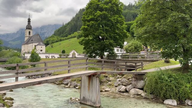 Mountain river, spring runs through Ramsau bei Berchtesgaden town. Wooden pedestrian bridge above. Parish Church of St. Sebastian (Pfarrkirche St. Sebastian Ramsau) in the backgroud. Ramsau bei Bercht
