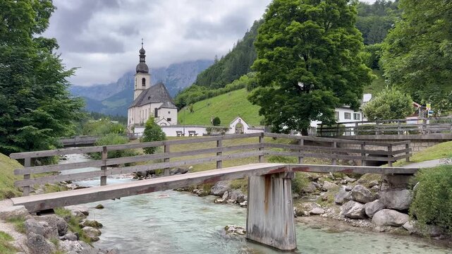 Mountain river, spring runs through Ramsau bei Berchtesgaden town. Wooden pedestrian bridge above. Parish Church of St. Sebastian (Pfarrkirche St. Sebastian Ramsau) in the backgroud. Ramsau bei Bercht