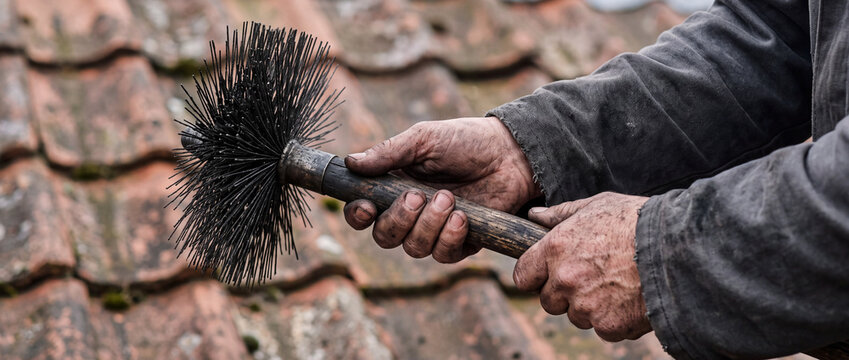 Chimney sweep holding brush on tiled roof showing weathered hands and traditional tool