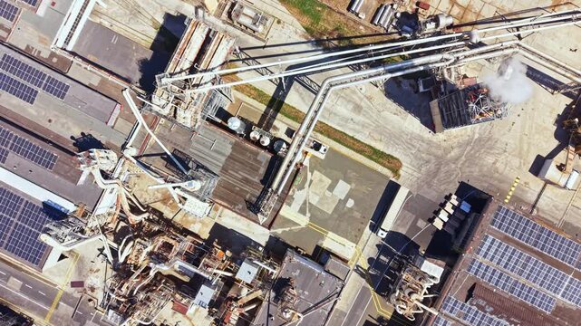 Pipes and structures fill the industrial site with solar panels on the rooftops. Smoke rises from one area as machinery operates under clear skies.