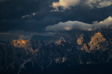 A dramatic mountain landscape showcasing golden sunlight hitting rugged peaks against a backdrop of dark stormy clouds. High contrast imagery perfect for travel and outdoor adventure themes. © marcodenaro