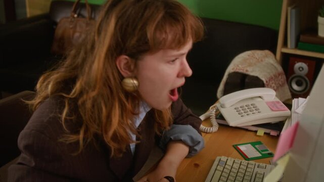 Young adult Caucasian woman yawning, rubbing eyes at retro computer desk, then answering landline phone. Perfect for 90s nostalgia marketing or vintage workplace concepts